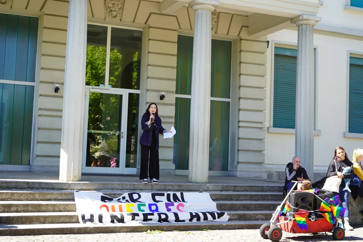 Man sieht eine Frau auf einer Treppe, die eine Rede hält. Vor ihr liegt ein Transparent.